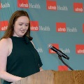 Individual stands at a wooden podium reading from notes during an indoor ceremony, speaking into a microphone, with a UTMB Health branded backdrop visible behind in a softly lit event space.
