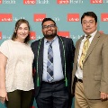 Three individuals pose indoors in front of a UTMB Health and John Sealy School of Medicine branded backdrop with repeating logos, dressed in semi-formal attire and standing close together for a group photo.