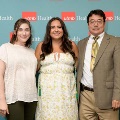 Three individuals pose indoors in front of a UTMB Health and John Sealy School of Medicine branded backdrop, with red logo panels, one wearing green honor cords, all dressed in semi-formal attire and standing close together for a photo.