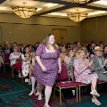 Individual walks down an aisle during an indoor ceremony while seated attendees on both sides applaud, with rows of chairs, patterned carpet, and softly lit chandeliers visible in the event space.