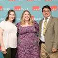 Three individuals pose indoors in front of a UTMB Health and John Sealy School of Medicine branded backdrop, with one wearing green honor cords, all dressed in semi-formal attire and standing close together for a photo.