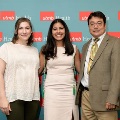 Three individuals pose indoors in front of a UTMB Health and John Sealy School of Medicine branded backdrop with repeating logos, one wearing green honor cords, all dressed in semi-formal attire and standing close together.