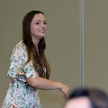 Individual in a light-colored floral dress stands among seated attendees during an indoor ceremony, with rows of chairs and a neutral wall in a softly lit event space visible in the background.