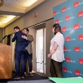 Two individuals embrace near a podium on a small stage during an indoor ceremony, with a UTMB Health branded backdrop to the side, another person holding green honor cords, and event lighting overhead.
