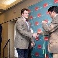 Two individuals shake hands on stage as one holds green honor cords, with a UTMB Health branded backdrop behind them and another person standing at a podium nearby in a formal indoor ceremony setting.
