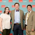 Three individuals pose indoors in front of a UTMB Health and John Sealy School of Medicine branded backdrop, with one wearing green honor cords, all dressed in semi-formal attire and standing close together for a photo.