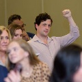 Audience members sit indoors during a ceremony, with one individual in the middle raising a hand while others look on, in a softly lit event space with rows of chairs and attendees seated closely together.