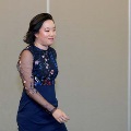 Individual in a navy blue dress with sheer embroidered sleeves walks indoors during an induction ceremony, with a neutral wall and softly lit event space visible in the background.