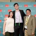 Three individuals pose indoors in front of a UTMB Health and John Sealy School of Medicine branded backdrop, with one wearing green honor cords, all in semi-formal attire and standing close together for a photo.