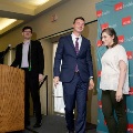 Individual wearing a suit and green honor cords walks off a stage near a podium in front of a UTMB Health branded backdrop, accompanied by another individual, with a speaker and microphone stand visible nearby.