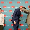 One individual places green honor cords over another individual’s head in front of a UTMB Health branded backdrop, while a third person stands nearby observing, all dressed in semi-formal attire.