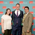 Three individuals pose indoors in front of a UTMB Health branded backdrop with repeating “utmb Health” logos, one wearing green honor cords, all dressed in semi-formal attire.