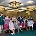 Multiple individuals stand in rows indoors raising one hand while holding printed programs during an oath ceremony, several wearing green honor cords, with seated attendees and a well-lit event space visible in the background.