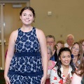 Individual in a sleeveless blue patterned dress stands in an indoor ceremony space while attendees seated behind clap, with double doors and rows of chairs visible in the softly lit background.