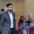 Individual in a suit walks forward during an indoor ceremony as seated attendees behind clap, with double doors and a softly lit event space visible in the background.