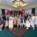 Large group of individuals pose indoors in front of a UTMB Health and John Sealy School of Medicine backdrop, framed by red, white, and gray balloon arches, with some seated in front and others standing behind in a well-lit event space.