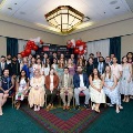 Large group of individuals pose indoors in front of a UTMB Health and John Sealy School of Medicine backdrop, framed by red, white, and gray balloon arches, with some seated in front and others standing behind in a well-lit event space.