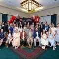 Large group of individuals pose indoors in front of a UTMB Health and John Sealy School of Medicine backdrop, framed by red, white, and gray balloon arches, with some seated in front and others standing behind in a well-lit event space.