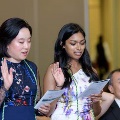 Two individuals stand indoors raising one hand while reading printed programs during an oath ceremony, wearing green honor cords, with additional participants and a seated audience visible in a softly lit event space.