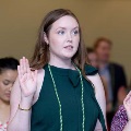 Individual wearing green honor cords stands indoors raising one hand during an oath-taking ceremony, while others nearby do the same, with a softly lit event space and seated audience visible in the background.