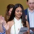 Individual wearing green honor cords stands indoors raising one hand while reading from a printed program during an oath ceremony, with other participants doing the same and a seated audience visible in the background.