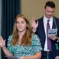 Two individuals stand indoors raising one hand while holding printed programs during an oath ceremony, wearing green honor cords, with other participants and a seated audience visible in a softly lit event space.