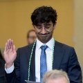Individual wearing a suit and glasses with green honor cords stands indoors raising one hand during an oath ceremony while holding a printed program, with other participants and a seated audience visible in the background.