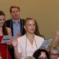Several individuals stand indoors raising one hand while holding printed programs during an oath-taking ceremony, wearing green honor cords, with a seated audience visible in the foreground and a neutral event space in the background.