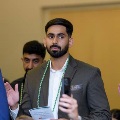 Individual wearing a suit and green honor cords stands indoors raising one hand during an oath ceremony while holding a program, with other participants and a seated audience visible in a softly lit setting.