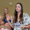 Individual stands indoors raising one hand while holding a printed program during an oath ceremony, wearing green honor cords, with other participants and a seated audience visible in the softly lit background.