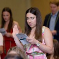 Individual stands indoors raising one hand while reading from a printed program during an oath ceremony, wearing green honor cords, with other participants and a seated audience visible in the background.
