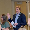 Several individuals stand indoors raising one hand during an oath-taking ceremony, one holding a program and wearing green honor cords, with double doors and a seated audience visible in the background.