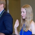 Individual in a blue outfit raises one hand during an oath-taking moment while another person stands nearby wearing honor cords, in an indoor ceremony setting with a neutral background.