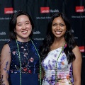 Two individuals pose indoors in front of a UTMB Health and John Sealy School of Medicine branded backdrop, framed by red and gray balloon decorations, with both wearing green honor cords and semi-formal attire.