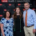 Three individuals pose indoors in front of a UTMB Health and John Sealy School of Medicine branded backdrop, framed by red, white, and gray balloon decorations, with one wearing green honor cords and all in semi-formal attire.