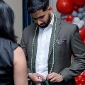 One individual stands indoors wearing a suit while another person places green honor cords around the neck, with red, white, and gray balloon decorations and a UTMB Health event backdrop visible in the background. Provide your feedback on BizChat