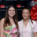 Two individuals pose indoors in front of a UTMB Health and John Sealy School of Medicine branded backdrop, with red, white, and gray balloon decorations on the side, one wearing green honor cords and both in semi-formal attire.