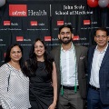 Four individuals pose indoors in front of a UTMB Health and John Sealy School of Medicine branded backdrop, framed by red, white, and gray balloon decorations, with one wearing honor cords and all in semi-formal attire.