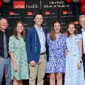 Seven individuals pose indoors in front of a UTMB Health and John Sealy School of Medicine backdrop, framed by red, white, and gray balloon decorations, with several wearing honor cords and semi-formal attire.