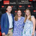 Three individuals pose indoors in front of a UTMB Health and John Sealy School of Medicine branded backdrop, framed by red, white, and gray balloon decorations, with two wearing honor cords and semi-formal attire.