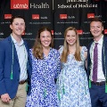 Four individuals pose indoors in front of a UTMB Health and John Sealy School of Medicine branded backdrop, framed by red, white, and gray balloon decorations, with several wearing honor cords and semi-formal attire.