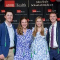 Four individuals pose indoors in front of a UTMB Health and John Sealy School of Medicine branded backdrop, framed by red, white, and gray balloon decorations, with several wearing honor cords and semi-formal attire.
