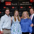 Four individuals pose indoors in front of a UTMB Health and John Sealy School of Medicine backdrop, framed by red, white, and gray balloons, with one wearing honor cords and others in semi-formal attire.