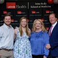 Four individuals pose indoors in front of a UTMB Health and John Sealy School of Medicine backdrop, framed by red, white, and gray balloons, with one wearing honor cords and others in semi-formal attire.