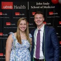 Two individuals pose indoors in front of a UTMB Health and John Sealy School of Medicine branded backdrop, wearing semi-formal attire, with red, white, and gray balloon decorations framing the sides.
