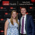 Two individuals pose indoors in front of a black UTMB Health and John Sealy School of Medicine backdrop, wearing semi-formal attire, with red, white, and gray balloon decorations framing the sides.