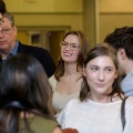 Several attendees stand indoors in a small group, engaged in conversation, wearing semi-formal attire in a softly lit reception space with others and doorways visible in the background.