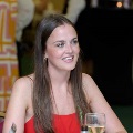 Individual seated at a table in a reception setting with a glass in front, wearing a red outfit, with other attendees and event decor visible in a softly lit background.
