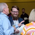 Three attendees stand indoors conversing, one holding a glass and papers, in a softly lit reception setting with other guests in the background wearing semi-formal attire.