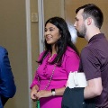 Three attendees stand indoors in conversation near a doorway, one wearing a bright pink outfit with honor cords and another holding a folder, with a neutral wall and event setting in the background.